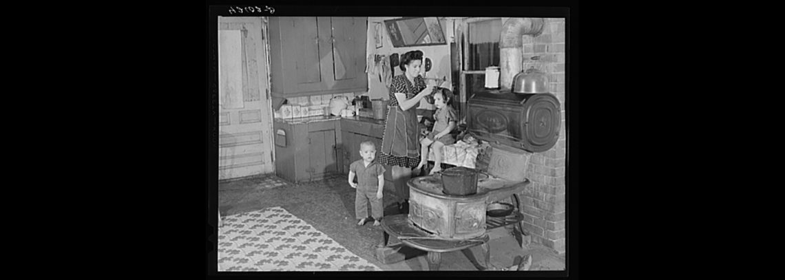 A black and white photograph of a woman and two small children in a kitchen around the 1930s.