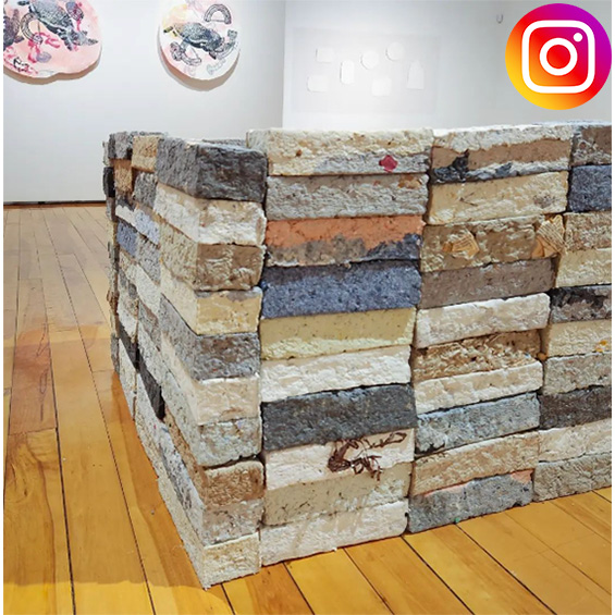 An image of a Gallery with wood floors and white walls with a sculpture iimmediately in the foreground resembling a cube of bricks. The Instagram logo is int he corner