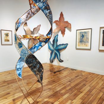 Center front: Kate Chenney Chappell, "Mother Ocean," 2021. Steel rod, printmaking (collagraph/monoprint) on paper, acrylic copper paint) 11 ft. 4 in. x 17 in. wide, on spiral (double helix). "Hidden Stories," 2023. University of Southern Maine Art Gallery. Photo by Kat Zagaria Buckley. 