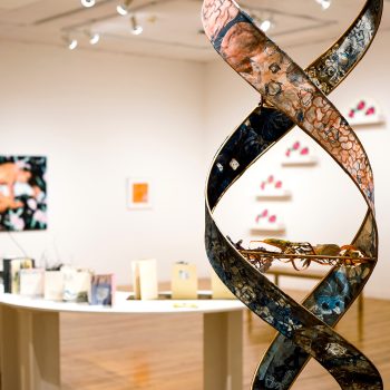 Front right: Kate Chenney Chappell, "Mother Ocean," 2021. Steel rod, printmaking (collagraph/monoprint) on paper, acrylic copper paint) 11 ft. 4 in. x 17 in. wide, on spiral (double helix). "Hidden Stories," Installation view, 2023. University of Southern Maine Art Gallery. Photo by Kat Zagaria Buckley. 