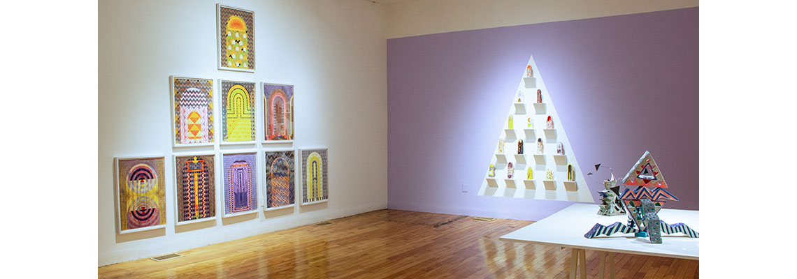 A gallery with wooden floors, one white wall, and one purple, ceramics are on a table in the foreground in abstract forms, and obelisk watercolors in a pyramid on a wall at left.
