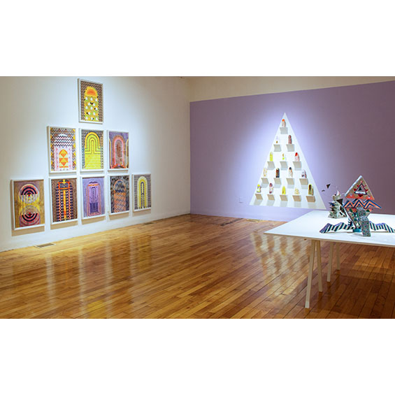 A gallery with wooden floors, one white wall, and one purple, ceramics are on a table in the foreground in abstract forms, and obelisk watercolors in a pyramid on a wall at left.