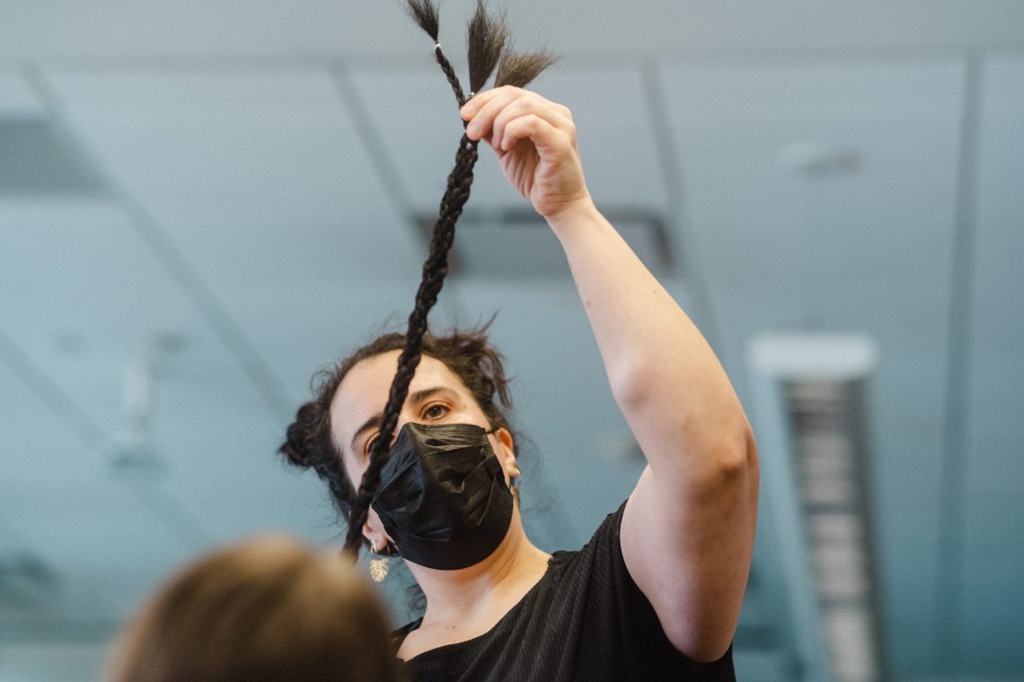 A woman with hair in alien buns holds  a braid of artificaal hair from end to end. She is wearing aa black surgical mask  and a black shirt.