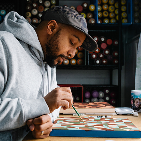 An artist works on a painting at a desk. In the background, neatly arranged spray paint cans with their nozels pointed towards the viewer.