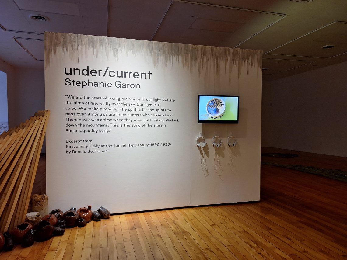 Stephanie Garon, "under/current," 2024. Installation, dimensions variable. Right side; Stephanie Garon, "Scattered Trees of a Storm-Swept Plain," 2024. Single channel video, running time 8 min 24 sec. Audio features Lucy Lippard, writer, activist, curator; Dwayne Tomah, Director of the Wabanaki Museum and Historic Preservationist of the Passamaquoddy Tribe; Severine Von Tscharner Fleming Welcome, Founder and Director of Smithereen Farms; Margaret Struzik, Pembroke Town Clerk; Mali Obomsawin, Musical artist; basket by: Passamaquoddy weaver Deborah Brooks of Sweet Grass Basketry. Filmed at Sipiyak/Pleasant Point and Indian Reservation, Maine, Potomac River. Installation view,"under/current" exhibition, University of Southern Maine Art Gallery, 2024. Photo: John Harlow.