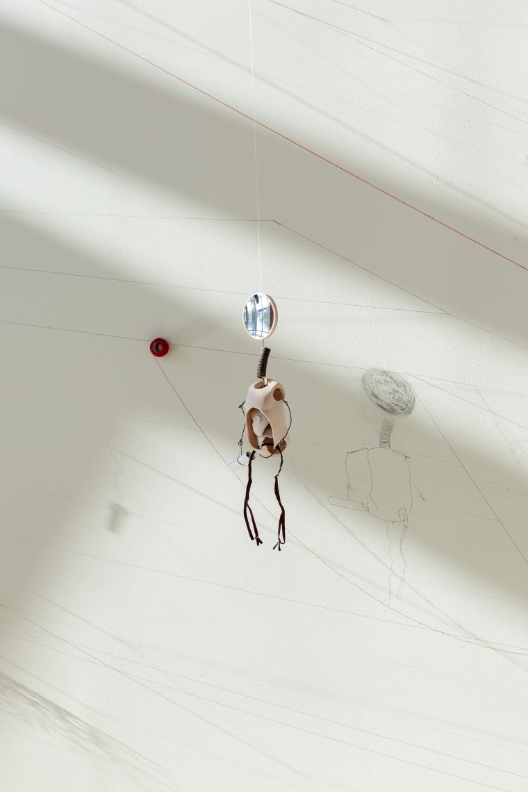 Work by Kristin Mariani. Background: "Leeward Ascent" (detail), 2026. Site specific wall installation: needles, thread, pencil, eraser, and silk remnant; 10 ft. high x 19 ft. wide x 16 in. deep. Atop Leeward Ascent: "Sibylle Helles (from Hellespontica)," 2025. Ceramic doll parts, magnifying mirror, found metal, shoulder straps, cardboard tubes, Post Its from Department of Reflection, thread, tape; 19 in. long. Part of "Of Sibyls and Source: Ranges and Ruptures," 2026. Crewe Center for the Arts at the University of Southern Maine. Photo: Marion Earley.