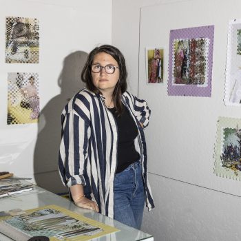 A woman with glasses and a striped shirt stands in a studio. Artwork is on the walls and she rests her hand on a glass table.