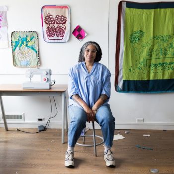 A woman in a blue shirt and jeans sits on a stool in a studio. Behind her are tapestries and a sewing machine.