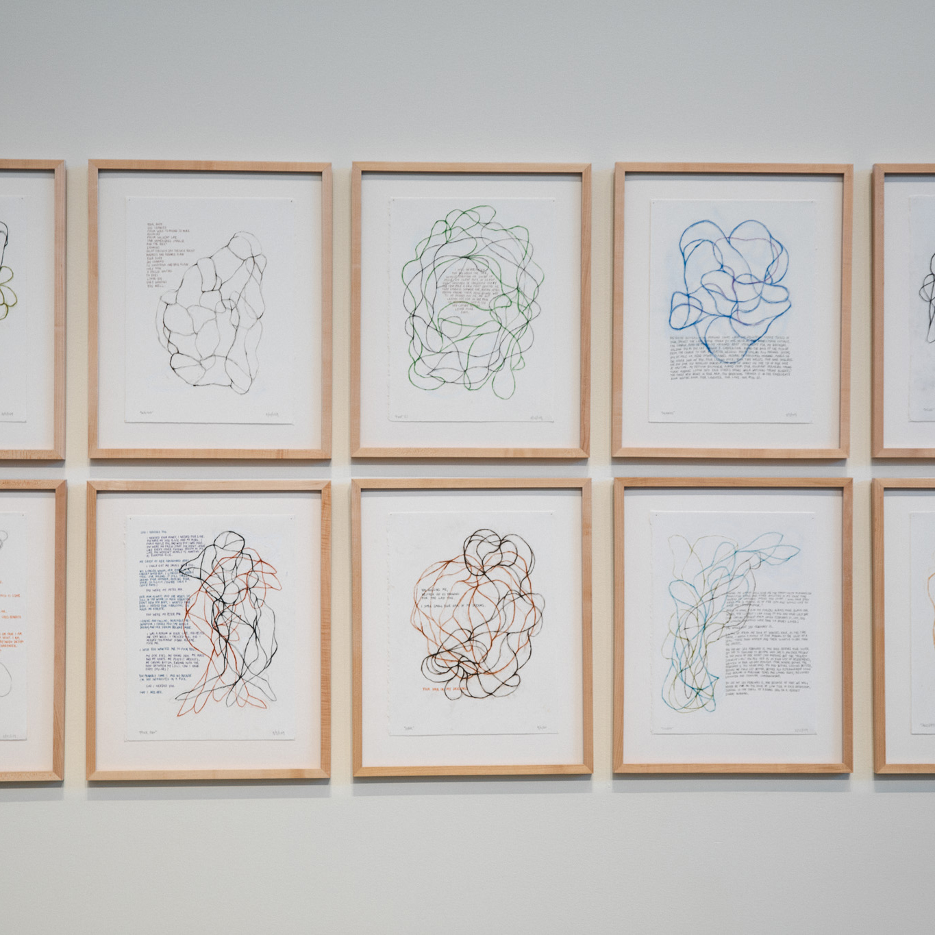 Two rows of artwork by Peter Bruun hang on a gallery wall in the Crewe Center for the Arts. Each artwork depicts an abstract line drawing with text and each piece is framed in a light wooden frame. 