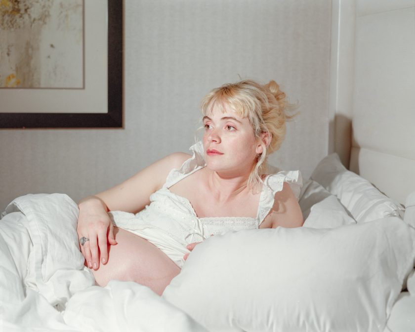 An image of a woman reclining on a bed with white sheets. The woman is pregnant, white, with blonde hair, wearing a white shirt. Her belly is protruting.
