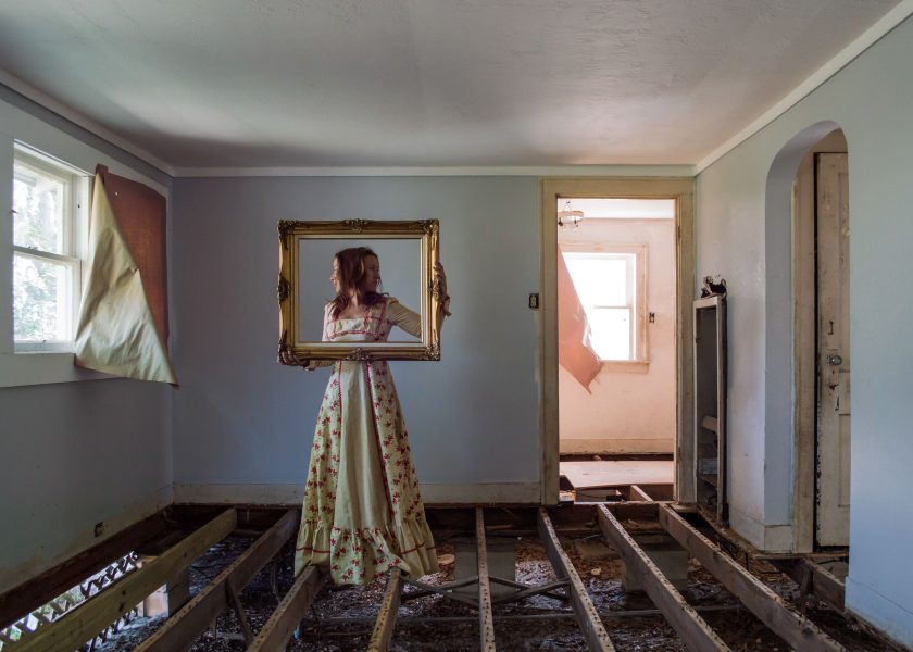 An image of a side profile of a woman in a dress standing on beams in a room without a solid floor. There is a doorway to her right and a window through it. The woman is holding a gold frame horizontally around her face.
