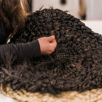 A long white table with braided artifical hair in blonde and black is laid out. An arm with a long black sleeve reaches out to continue braiding. The braids are in circles, like trivets.