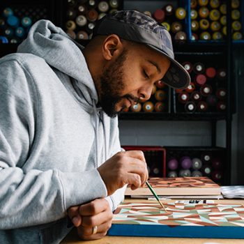 A man with a beard, a cap, and a grey hoodie paints at a desk.