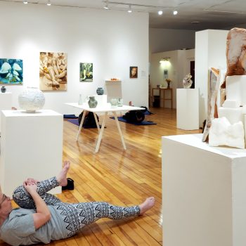 A white walled gallery with wooden floors. Attendees engage in an exercise on yoga mats surrounded by hanging artworks and sculptures on pedastals.