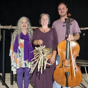 Three individuals stand together. The one on the left is a woman with long hair dresse din purple, in the center, a woman in a mauve dress holding a mobile, and at right, a tall man in a lighter mauve shirt holding a cello and bow.