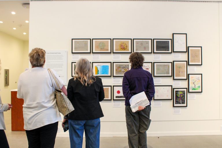 A group of three people stand in front a series of small, framed artworks in the Crewe Center for the Arts. The observers face the artwork hung on the gallery wall, their backs are turned toward the camera. The atmosphere is bright and pensive. The artwork depict colorful drawings of symbols and words.