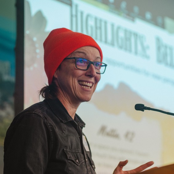 a student wearing an orange hat is smiling, speaking into a microphone, with a slide presentation displayed in the background