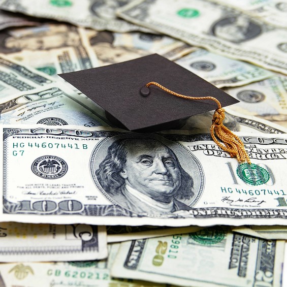 a black graduate cap with gold tassel sits atop a $100 dollar bill, as well as other dollar bills of various denominations