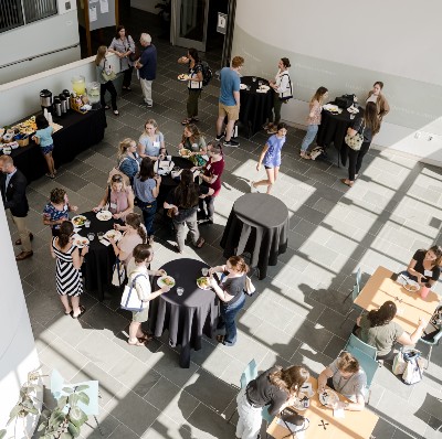 An arial view of an event. A sunny room has a buffet table of food in the upper left, while people mingle around bistro tables throughout the rest of the frame