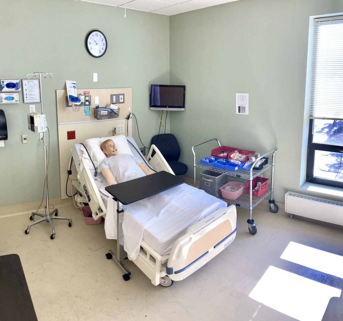 A patient mannequin sits in a hospital bed. A tray rests over the bottom half of the bed. A monitor, and many other medical devices sit to the sides and above the bed.