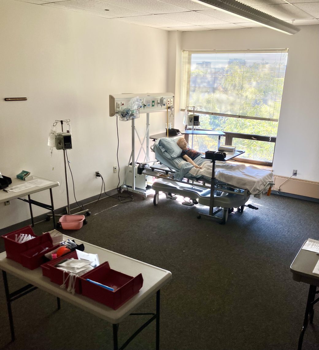 A patient mannequin sits in a hospital bed. A tray rests over the bottom half of the bed. Several medical devices sit to the sides of and above the bed. In the foreground is a table with several red bins, each containing items, such as bags and syringes.