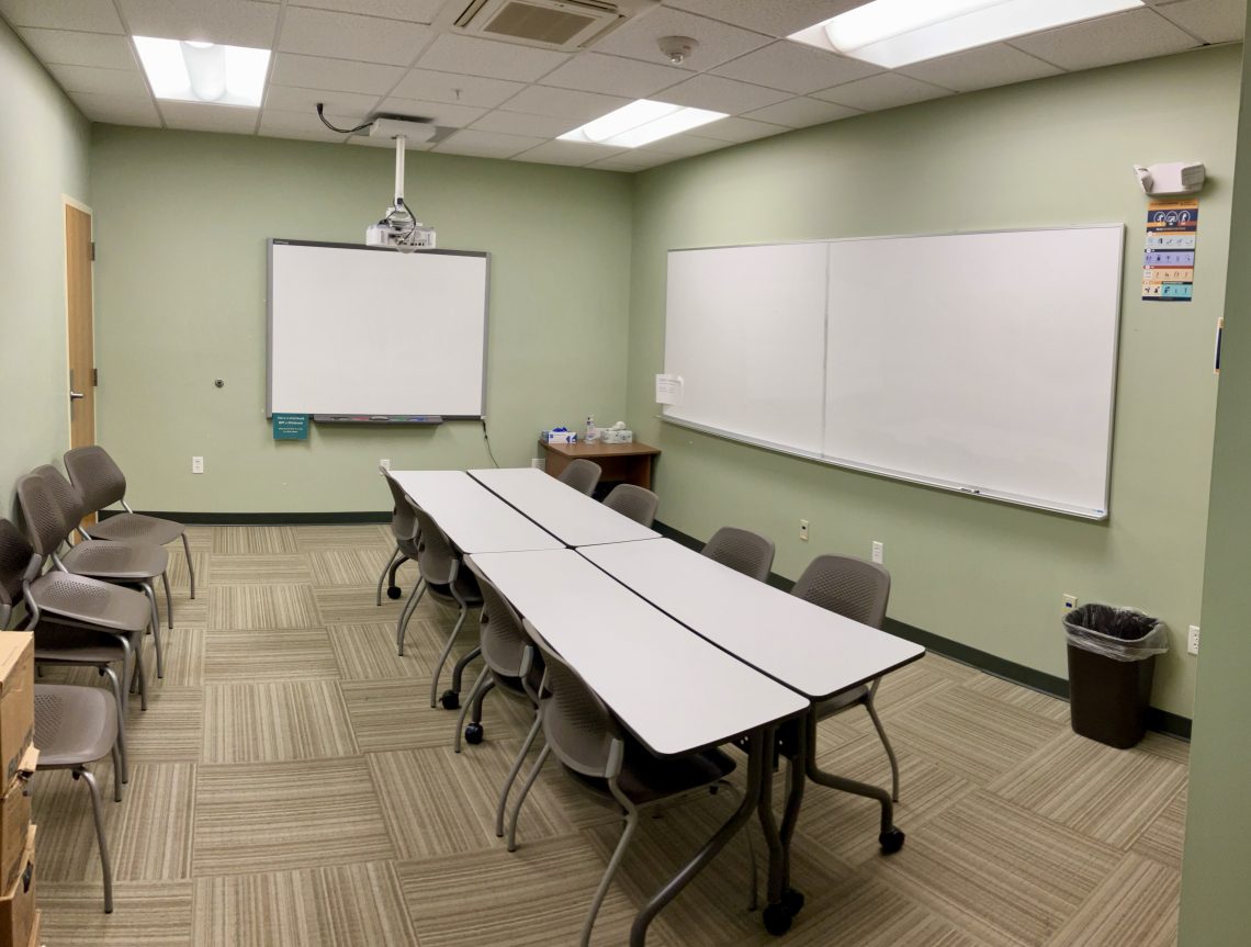 A classroom with green walls, tan, patterned carpet, two white boards on one wall, and a projector screen on the far wall. A table with several chairs at it rests in the center of the room. To the left, more chairs sit lined up against the wall.