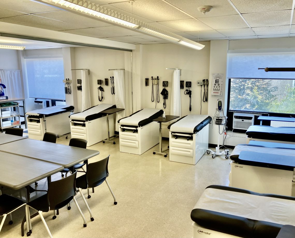 A wall with four medical examination beds covered in sanitary paper. To the right, on another wall, are three more beds. In the foreground, several tables are pushed together to make one large seating area surrounded by chairs. Two large windows are in the background.