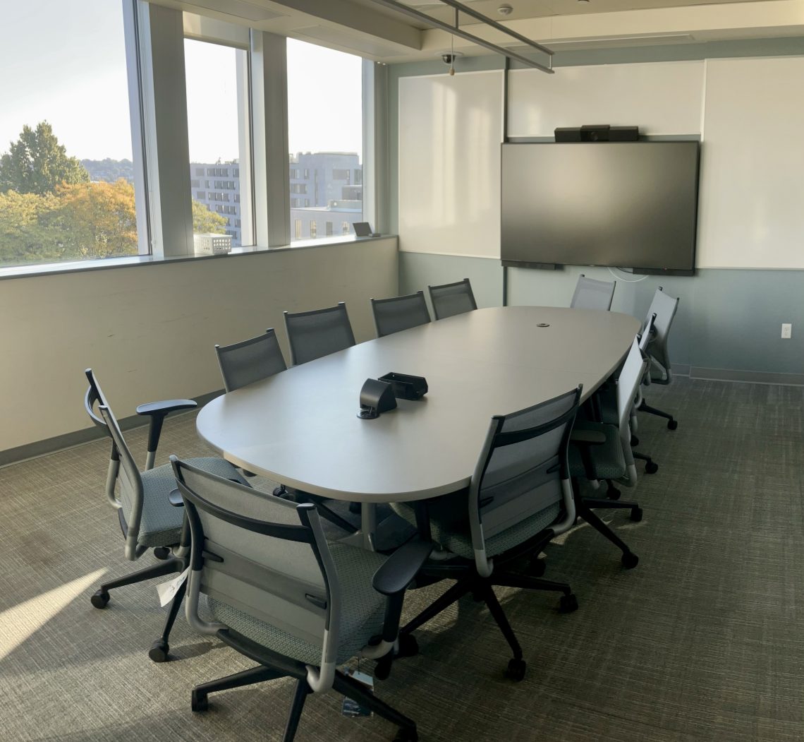 A conference room with a camera and microphone hanging from the ceiling, over a long table surrounded by about ten chairs. To the left are three open windows, and on the far wall is a tv with its own camera.