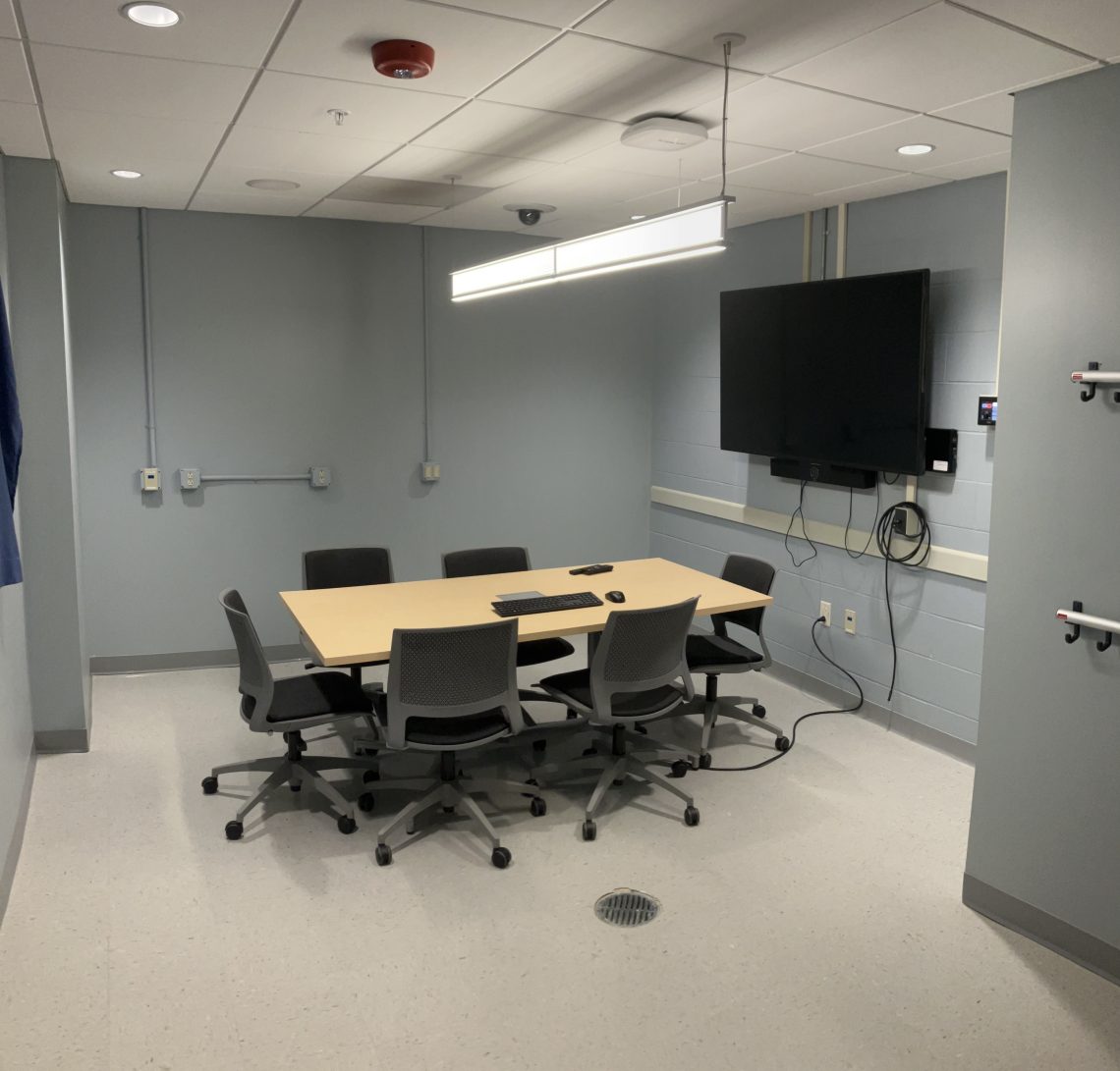 An observation room with a table and six chairs, a television, and wires connected to the equipment.