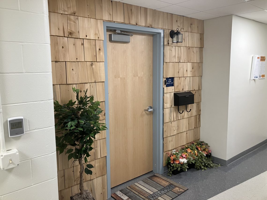 A door inside an academic building hallway that is made to look like the door to a patient's house. Un-stained paneled siding covers the wall around the door. Near the door is a black mailbox on the wall. A welcome mat sits in front of the door. On either side of the door are decorative flowers and trees.