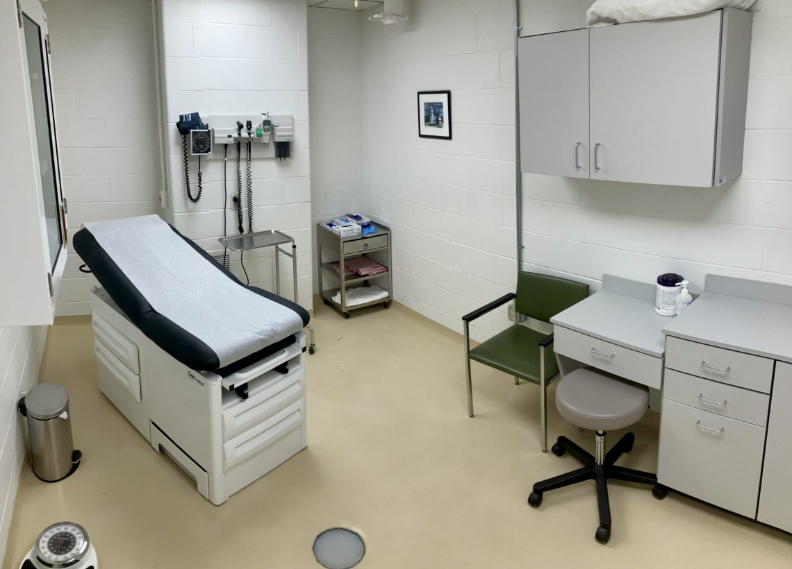 A simulation patient room. One medical bed sits in the room. On the right side are a chair, a stool, and counter space with sanitizing products. On the far wall sits medical equipment. In front of the far wall sits a tray stand on wheels and a small cart on wheels which has blankets and medical gloves.