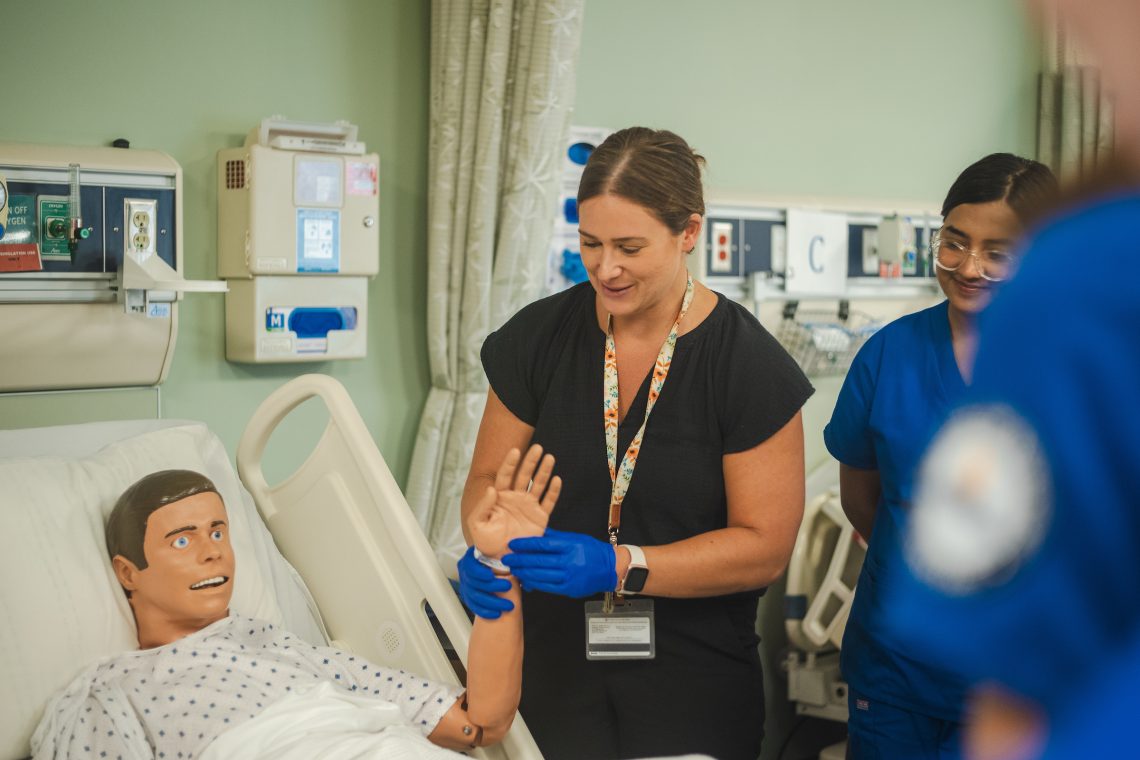 A faculty member stands next to a medical mannequin's bed, holding the mannequin's left hand up as if checking for a pulse. A student looks on from next to the faculty member. In the background on the wall is a number of pieces of medical equipment and a dispenser for medical gloves.