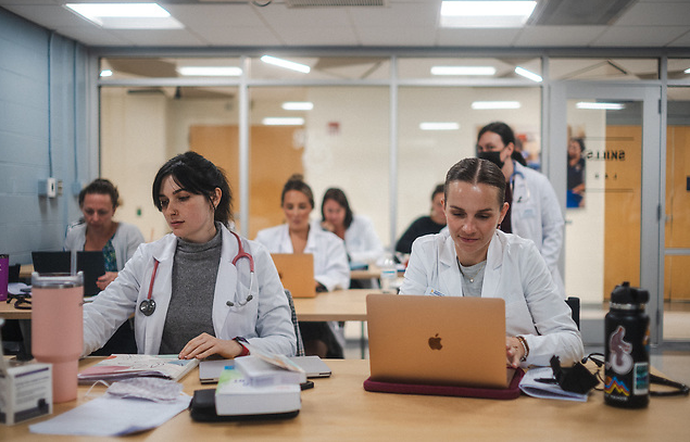Two students in lab coats sitting at a desk. One reads a computer screen and the other takes notes. Other students and a faculty member sit and stand in the background.
