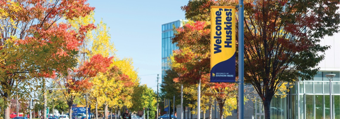 A student walking up Bedford St on a sunny autumn day. A lamppost bears a banner that reads 'Welcome Huskies' above the USM logo.