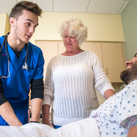 A student wearing School of Nursing scrubs checks on a patient while an instructor wearing a white sweater monitors the interaction.