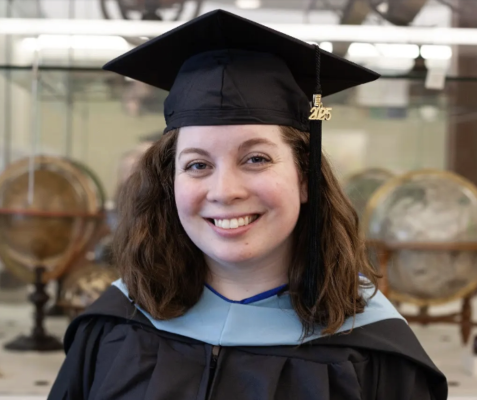 Smiling graduate wearing a cap and gown with a light blue hood, standing indoors in front of a display of antique globes
