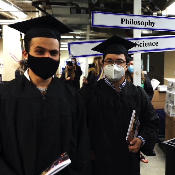 Two masked students in their graduation robes pose for the camera behind the scenes.