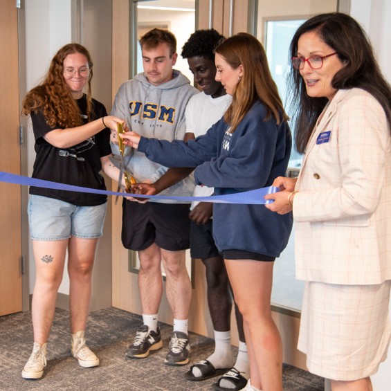 Four students combine to wield a pair of scissors while President Jacqueline Edmondson holds the ribbon at a ceremony to open the new Portland Commons dormitory.