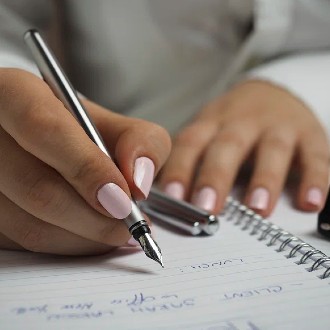 Hand with pink painted fingernails writing in a notebook with a pen.