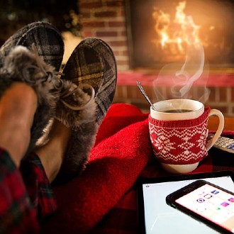 Slippered feet in front of a fireplace with a hot beverage in a mug, next to a cel phone and TV remote.