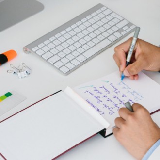 Person writing a to do list in a notebook in front of a computer