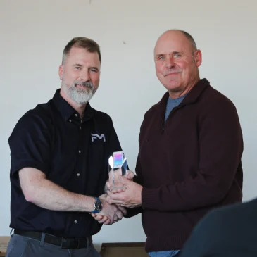 Daniel Lee of Factory Mutual shakes hands with Brian Bishop, USM’s assistant director of Facilities Management for Operations, as he presents a glass safety award. Both are smiling at the camera in a campus meeting room.