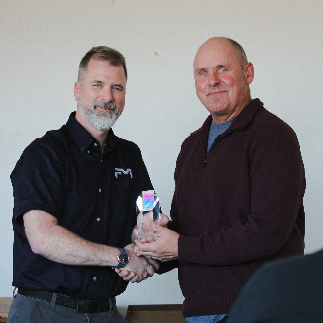 Daniel Lee of Factory Mutual shakes hands with Brian Bishop, USM’s assistant director of Facilities Management for Operations, as he presents a glass safety award. Both are smiling at the camera in a campus meeting room.