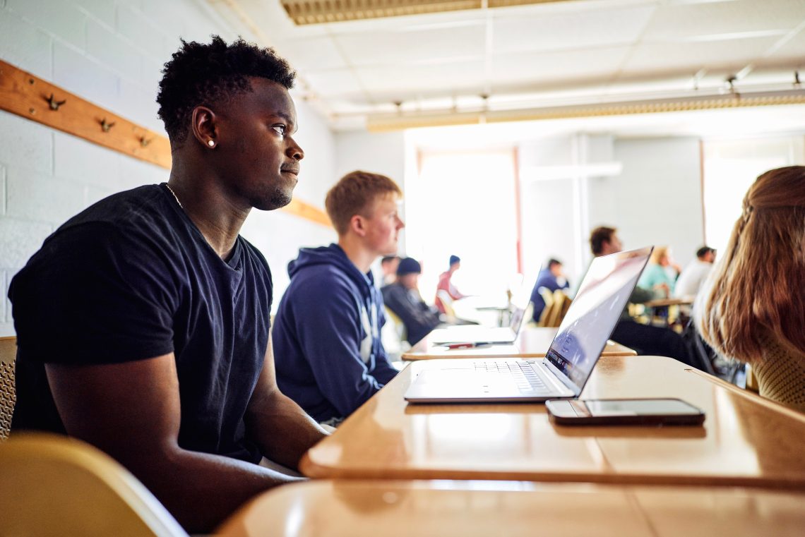 Student seated at a desk in a classroom, with natural light coming through windows in the background.