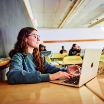 Student using a laptop in a classroom, seated in front of a chalkboard.