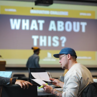 Student reviews challenge materials at What About This? Innovation Challenge kickoff event with "What About This" projected on screen in background