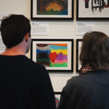 Two people examine a framed pastel drawing of a heart and rainbow, created during a community art workshop as part of USM's 'Even As We Grieve' exhibition at the Crewe Center for the Arts.
