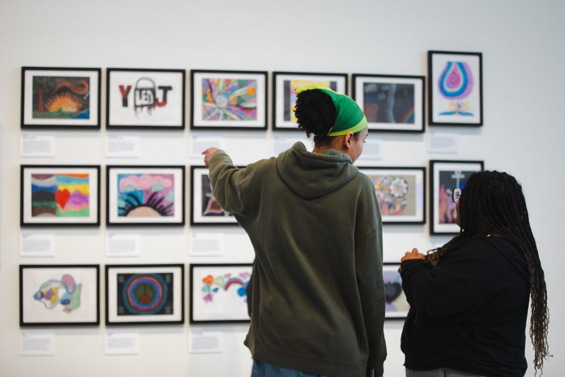 Two visitors point at framed artwork created by community members during nonprofit art workshops, on view as part of 'Even As We Grieve' at the Crewe Center for the Arts.