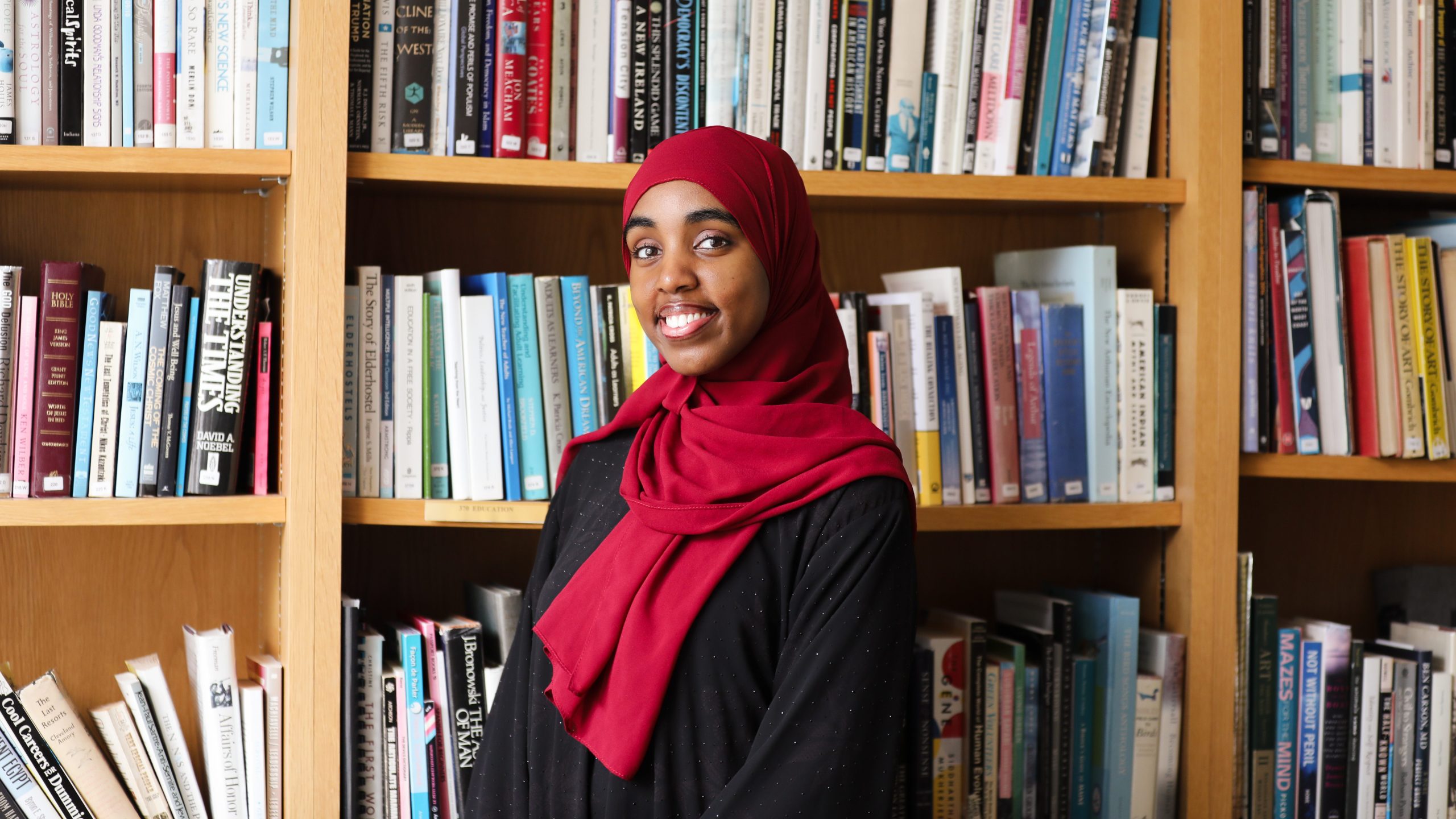 Najmo Ahmed, USM's 2026 student commencement speaker, smiles in front of a wall of bookshelves.
