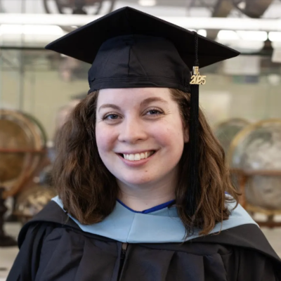 Smiling graduate wearing a cap and gown with a light blue hood, standing indoors in front of a display of antique globes